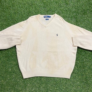 May include: A cream-colored v-neck sweater with a small polo player logo on the left chest.