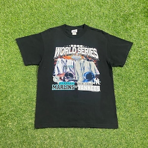 Puede incluir: Camiseta negra con un gráfico de la Serie Mundial de 2003 con los Marlins de Florida y los Yankees de Nueva York. El texto "2003 World Series" está impreso en blanco con un contorno negro. Los logotipos de los equipos son visibles en las camisetas.