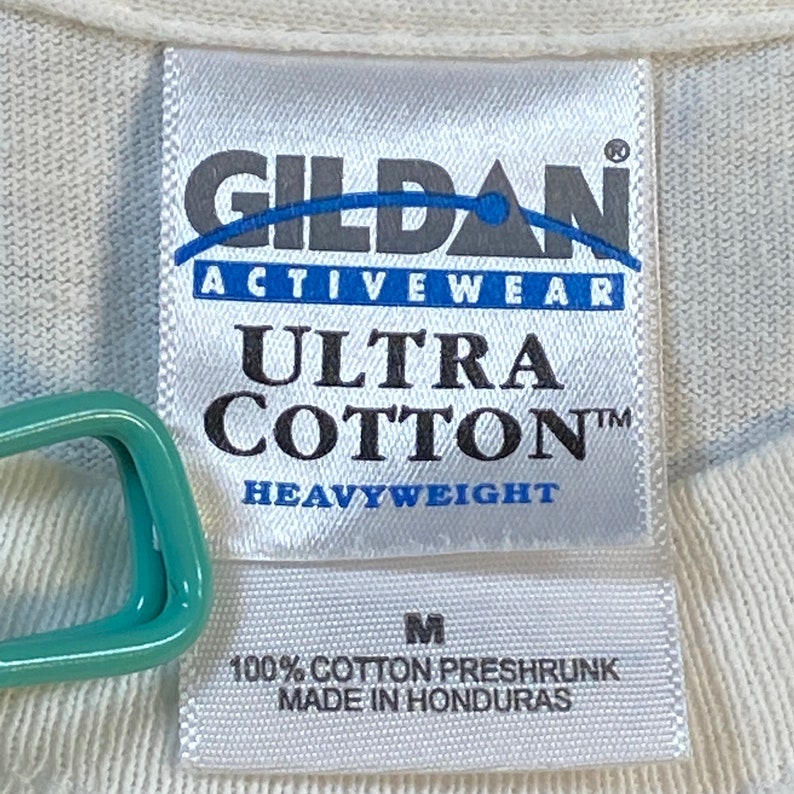 Może przedstawiać: Biała metka koszulki Gildan Ultra Cotton Heavyweight w rozmiarze M, 100% bawełna, wstępnie skurczona, wyprodukowana w Hondurasie.