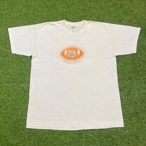 Puede incluir: Camiseta blanca con un gráfico de fútbol americano en naranja y marrón, y el texto "Penn State, Florida State". La camiseta está colocada sobre una superficie verde.