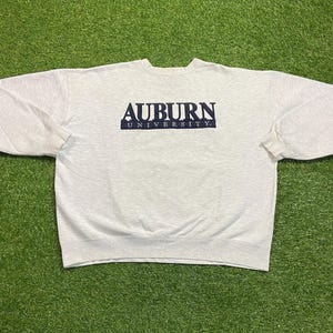 May include: A light gray sweatshirt with the text "AUBURN UNIVERSITY" in navy blue. The sweatshirt is made of a soft material and has long sleeves. The text is centered on the chest.