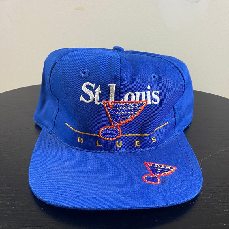 K&ouml;nnte beinhalten: Eine blaue Baseballkappe mit einem wei&szlig;en gestickten St. Louis Blues Logo, das eine Musiknote und einen Hockeyschl&auml;ger zeigt. Das Logo ist gold umrandet und das Wort "Blues" ist in Gold unter dem Logo geschrieben.