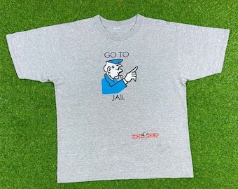May include: A gray t-shirt with a Monopoly game character illustration. The character is wearing a blue uniform and pointing with his right hand. The text "Go To Jail" is printed above the character and "MONOPOLY" is printed below the character.