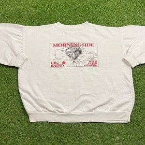 Peut inclure: Sweat-shirt blanc à col rond avec un motif de l'émission de radio CBC "Morningside with Peter Gzowski". Le motif comprend une carte et un portrait de Peter Gzowski en rouge et noir. Le sweat-shirt a des poignets et un ourlet côtelés.