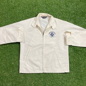 Puede incluir: Una chaqueta blanca con cremallera que tiene un parche bordado en azul que dice "Camp Staff" y un triángulo con un pino dentro. La chaqueta tiene dos bolsillos en la parte delantera.