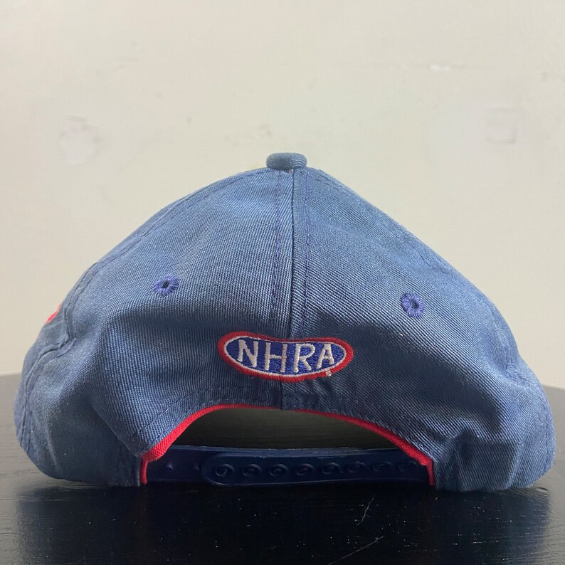 May include: A blue baseball cap with a red brim and the embroidered letters "NHRA" in white. The cap has a snapback closure and a small button on top. The hat is made of a durable fabric.