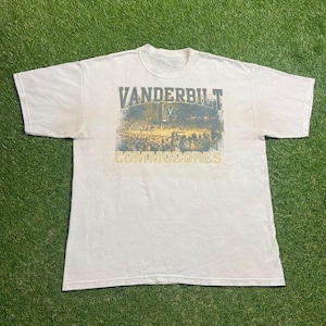 May include: Off-white t-shirt featuring the text "VANDERBILT COMMODORES" in a distressed font. The graphic includes a vintage-style illustration of a stadium scene. The shirt is made of a soft, comfortable material.