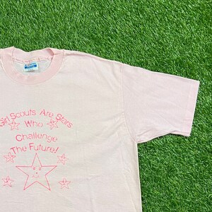 Puede incluir: Una camiseta rosa claro con cuello y mangas rosas. La camiseta presenta el texto "Girl Scouts Are Stars Who Challenge The Future!" en rosa, junto con gr&aacute;ficos de estrellas. La camiseta est&aacute; hecha de un material suave.