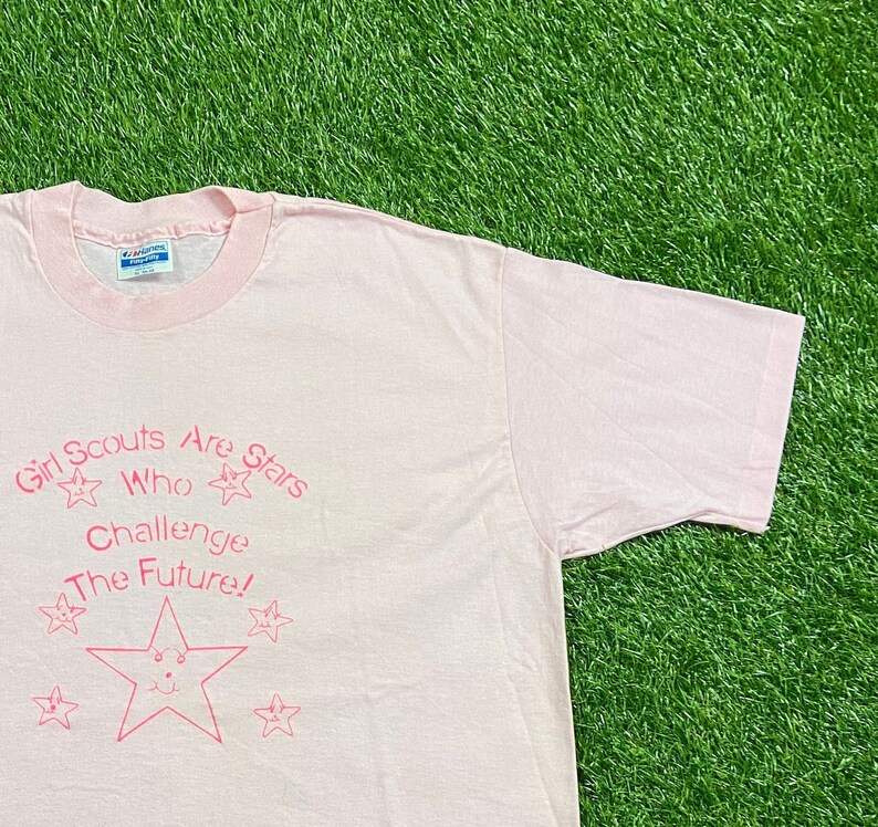 Puede incluir: Una camiseta rosa claro con cuello y mangas rosas. La camiseta presenta el texto "Girl Scouts Are Stars Who Challenge The Future!" en rosa, junto con gr&aacute;ficos de estrellas. La camiseta est&aacute; hecha de un material suave.