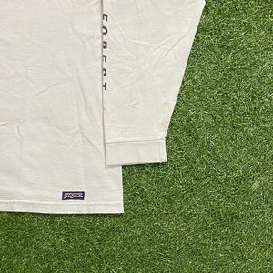 Może przedstawiać: Biała koszulka z długim rękawem z napisem "FOREST" wydrukowanym pionowo na rękawie w ciemnym kolorze. Koszulka ma logo "JANSPORT" w lewym dolnym rogu.