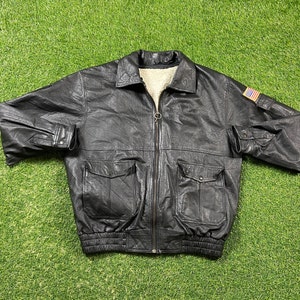 May include: A black leather bomber jacket with a zipper closure, two pockets, and a fur-lined collar. The jacket has a small American flag patch on the right sleeve.