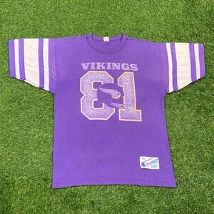 May include: Purple vintage t-shirt with the word "VIKINGS" and the number "81" in white and gold. The sleeves have white stripes. A Champion tag is visible. The shirt is on a green grass background.