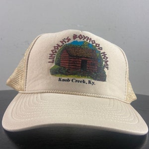 Puede incluir: Una gorra de camionero beige con una cabaña marrón y el texto "Lincoln's Boyhood Home Knob Creek, Ky." impreso en la parte delantera.