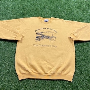 May include: A yellow sweatshirt with a graphic print of a town with the text "Home of the Rocket Boys The Coalwood Way".