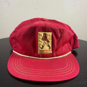 Op de afbeelding: Rode baseballpet met een witte touwrand en een Johnnie Walker Red-label met een man in een rood jasje die met een stok loopt.