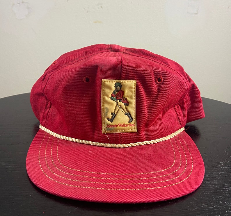 Op de afbeelding: Rode baseballpet met een witte touwrand en een Johnnie Walker Red-label met een man in een rood jasje die met een stok loopt.
