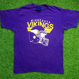 May include: Purple t-shirt with a yellow and white Minnesota Vikings logo. The logo features a football helmet and the words "Minnesota Vikings".