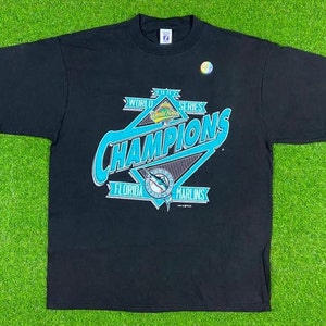 May include: Black t-shirt with a teal blue and white graphic of the 1997 World Series Champions Florida Marlins logo. The logo features a marlin fish, a baseball, and the words "World Series Champions Florida Marlins".