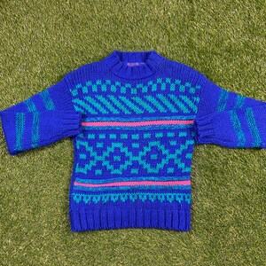 Peut inclure: Un pull en tricot bleu avec un motif géométrique turquoise et rose. Le pull a des manches longues et un col rond.