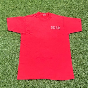 Peut inclure: Un t-shirt rouge vif à manches courtes. Le texte "SDSU" est imprimé en blanc sur le haut de la poitrine gauche. Le t-shirt est en matière souple et est probablement vintage.