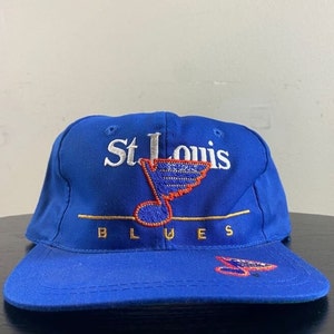 K&ouml;nnte beinhalten: Eine blaue Baseballkappe mit einem wei&szlig;en gestickten St. Louis Blues Logo, das eine Musiknote und einen Hockeyschl&auml;ger zeigt. Das Logo ist in Gold umrandet.