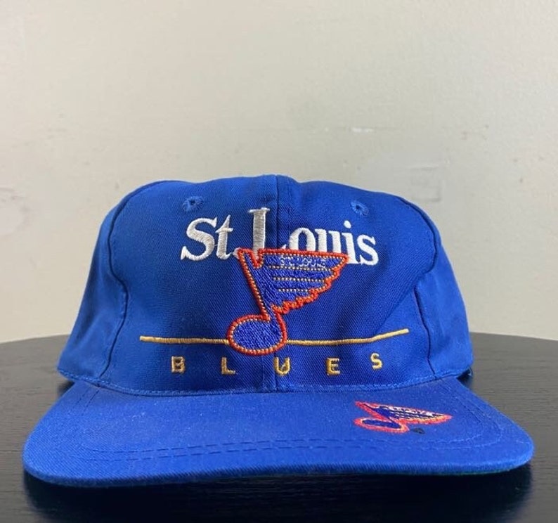 K&ouml;nnte beinhalten: Eine blaue Baseballkappe mit einem wei&szlig;en gestickten St. Louis Blues Logo, das eine Musiknote und einen Hockeyschl&auml;ger zeigt. Das Logo ist in Gold umrandet.