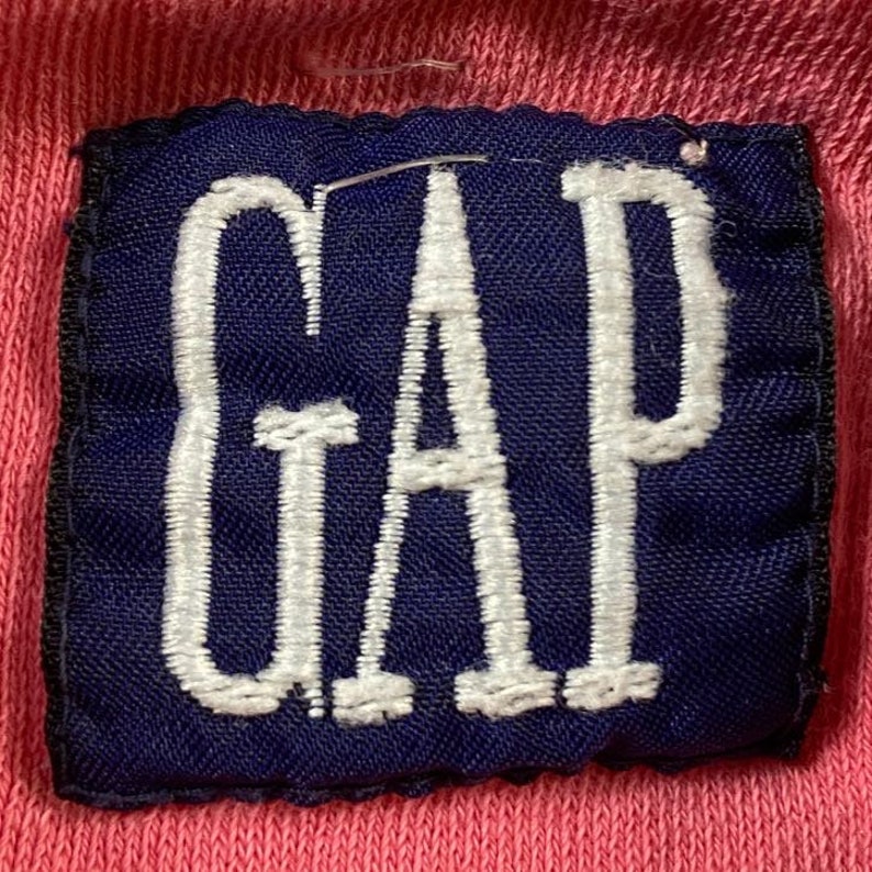 May include: A close-up of a blue and white embroidered patch with the word "GAP" in white lettering on a pink background.
