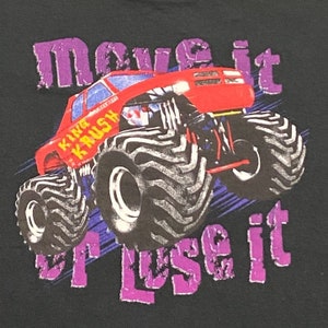 Może przedstawiać: Czarny T-shirt z grafiką czerwonego monster trucka. Ciężar&oacute;wka ma duże opony i napis "King Krush" na boku. Tekst "Move it or lose it" jest nadrukowany na koszulce na fioletowo.