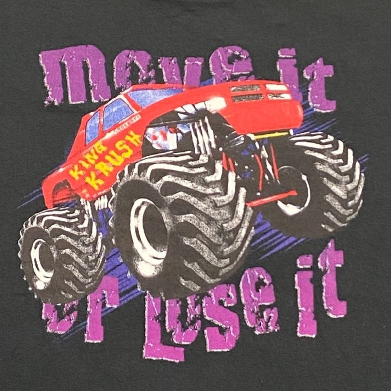 Może przedstawiać: Czarny T-shirt z grafiką czerwonego monster trucka. Ciężar&oacute;wka ma duże opony i napis "King Krush" na boku. Tekst "Move it or lose it" jest nadrukowany na koszulce na fioletowo.