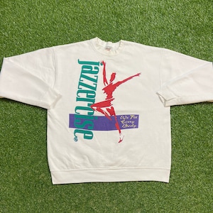 May include: White sweatshirt with a green and purple graphic of a dancer and the text "Jazzercise" with the tagline "We Fit Every Body".