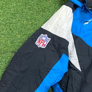 Peut inclure: Veste NFL noire, bleue et blanche avec le logo NFL sur la manche. La veste a une capuche bleue et un logo Reebok. La veste est faite d'un mat&eacute;riau brillant, possiblement en nylon.