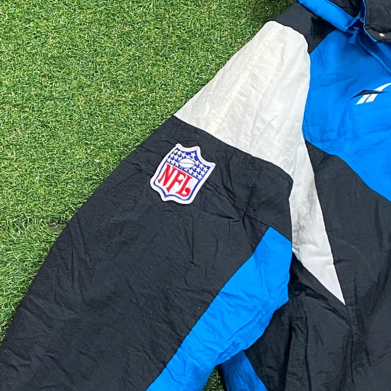 Peut inclure: Veste NFL noire, bleue et blanche avec le logo NFL sur la manche. La veste a une capuche bleue et un logo Reebok. La veste est faite d'un mat&eacute;riau brillant, possiblement en nylon.