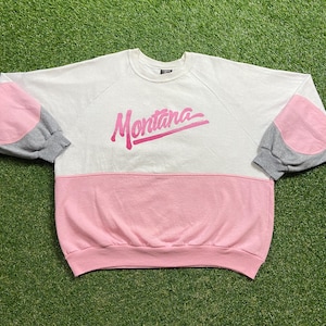 May include: A white, pink, and gray sweatshirt with the word "Montana" in pink script across the chest. The sleeves have gray cuffs and pink elbow patches. The bottom half of the sweatshirt is pink.