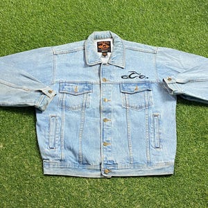 May include: Light blue denim jacket with a button-down front, two chest pockets, and two side pockets. Features a small logo patch on the collar and embroidered design on the chest. The jacket is laid flat on a green surface.
