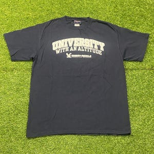 May include: Navy blue t-shirt with the text "UNIVERSITY WITH AN ALTITUDE" in white block letters. Below the text is a graphic of a bird and the text "EMBRY-RIDDLE AERONAUTICAL UNIVERSITY". The shirt is on a green surface.