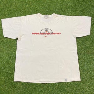 May include: Off-white t-shirt featuring the Manchester United logo. The logo includes a soccer ball graphic above the words "MANCHESTER UNITED" in red, with a small Nike swoosh below. The shirt is made of a soft material.