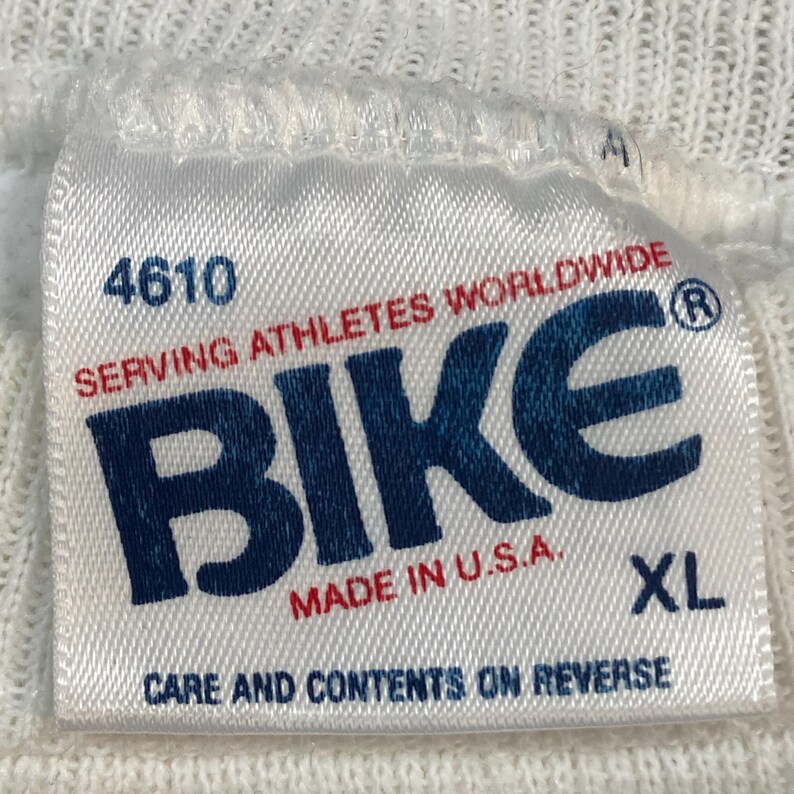 May include: White fabric tag with the word "BIKE" in navy blue, and the text "SERVING ATHLETES WORLDWIDE" in red. The tag also includes the text "MADE IN U.S.A." and "XL". The number "4610" is printed at the top of the tag.