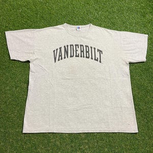May include: Light gray t-shirt with the word "VANDERBILT" arched across the chest in dark gray letters. The shirt is made of a soft material and has short sleeves. The shirt is laid flat on a green surface.