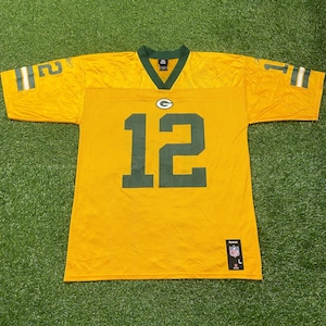 Puede incluir: Una camiseta de fútbol dorada y verde con el número 12 en verde en la parte delantera. La camiseta tiene detalles verdes en el cuello y las mangas. Los logotipos de la NFL y Reebok son visibles.