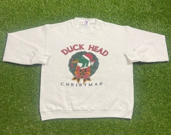 Può includere: Felpa bianca con una testa di anatra verde che indossa un cappello da Babbo Natale in una ghirlanda verde con un nastro rosso e il testo "Duck Head Christmas".