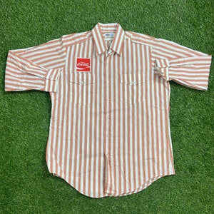 Può includere: Una camicia a maniche lunghe abbottonata a righe bianche e arancioni con una toppa rossa Coca-Cola sul petto sinistro. La camicia ha due tasche sul petto.