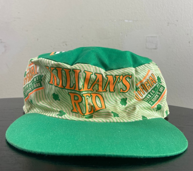 May include: Green and white baseball cap with the words "Killian's Red" in orange and yellow. The cap has a green brim and a white crown with a green and white pattern. The text "Official Dinner - St. Patrick's Day" is printed on the side of the cap.