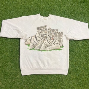 May include: White sweatshirt featuring a graphic of two white tigers resting on green grass. The tigers have black stripes and are in a relaxed pose. The sweatshirt has long sleeves and a crew neck.