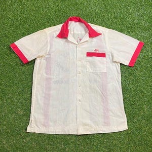 May include: Cream-colored button-down shirt with red collar, cuffs, and pocket trim. The shirt features the name "JIM" embroidered on the pocket. The shirt is short-sleeved and has a classic, vintage style.