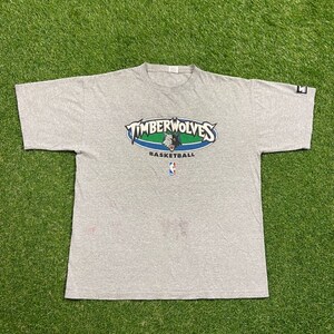 May include: A heather gray t-shirt featuring the TimberWolves basketball logo in blue, green, and white. The NBA logo is below the team name. The shirt is short-sleeved.