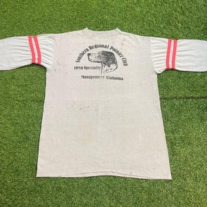 May include: A light gray t-shirt with red stripes on the sleeves. The shirt features a graphic of a dog and text that reads "Southern Regional Pointer Club 1986 Specialty Montgomery Alabama."