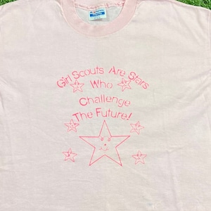 Puede incluir: Camiseta rosa claro con cuello rosa y texto rosa "Girl Scouts Are Stars Who Challenge The Future!". El dise&ntilde;o incluye estrellas.