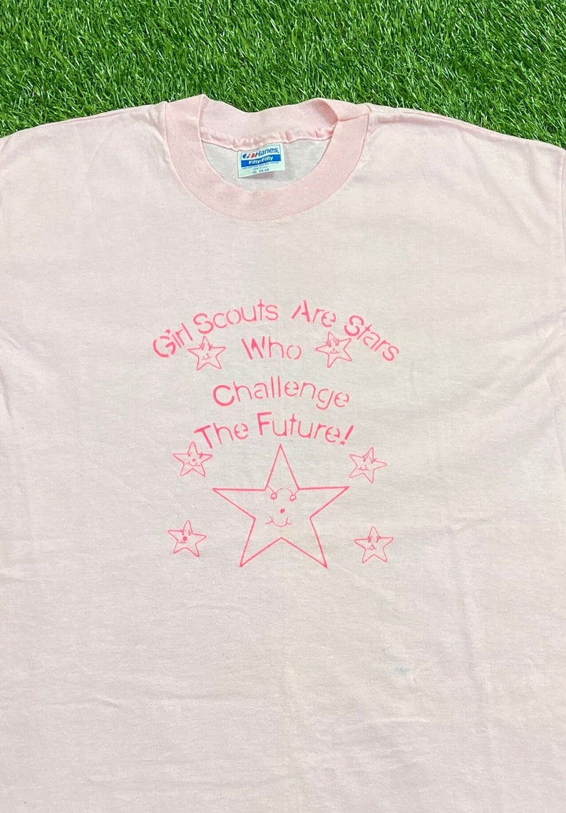 Puede incluir: Camiseta rosa claro con cuello rosa y texto rosa "Girl Scouts Are Stars Who Challenge The Future!". El dise&ntilde;o incluye estrellas.