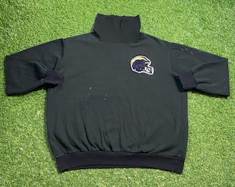 Puede incluir: Sudadera verde oscuro de cuello alto con un parche de casco de fútbol americano. La sudadera tiene puños negros y un dobladillo inferior negro. El parche del casco de fútbol es amarillo, negro y blanco.