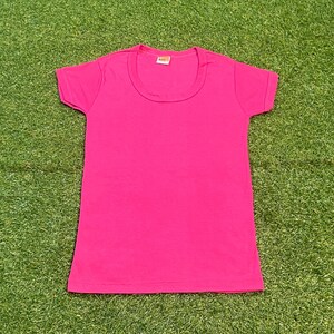 May include: A vibrant pink t-shirt with a scoop neck and short sleeves. The shirt is made of a soft material and is displayed on a green surface. The t-shirt is a solid color and has a simple, classic design.